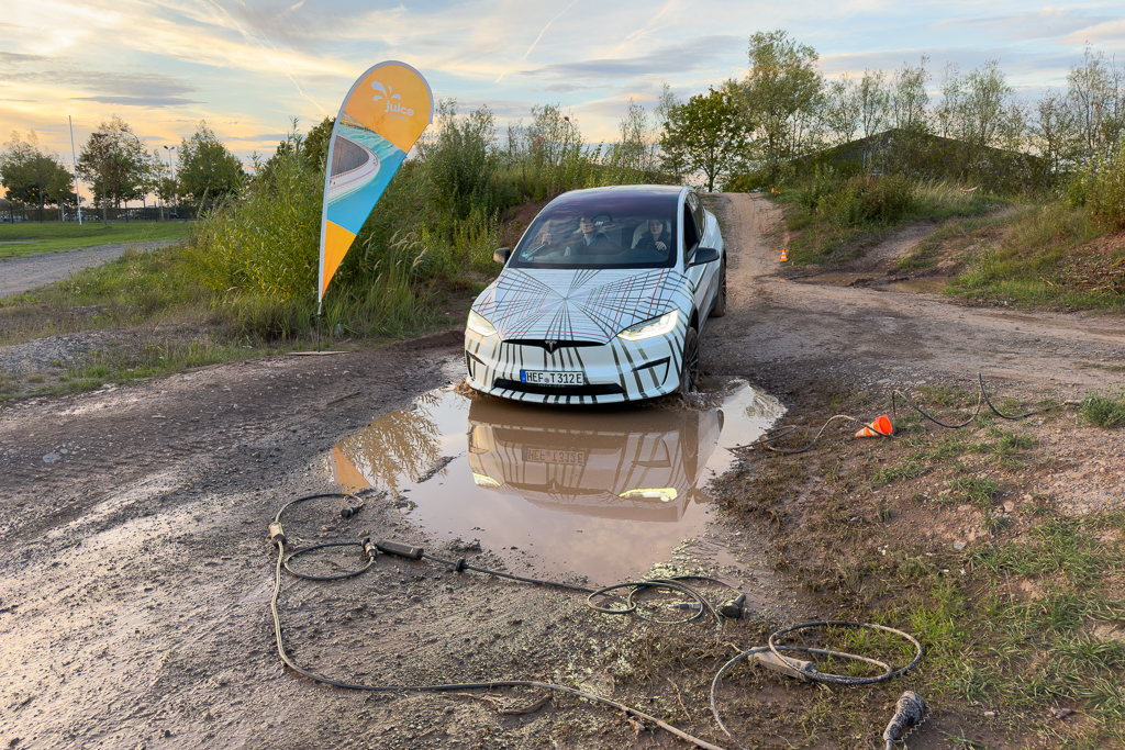 An electric vehicle partially submerged in a muddy puddle on a dirt path, with a Juice branding flag in the background.