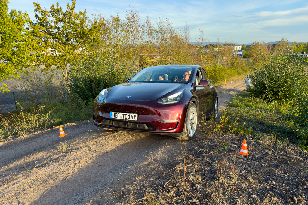 A maroon Tesla vehicle navigating a dirt path surrounded by greenery and cones.