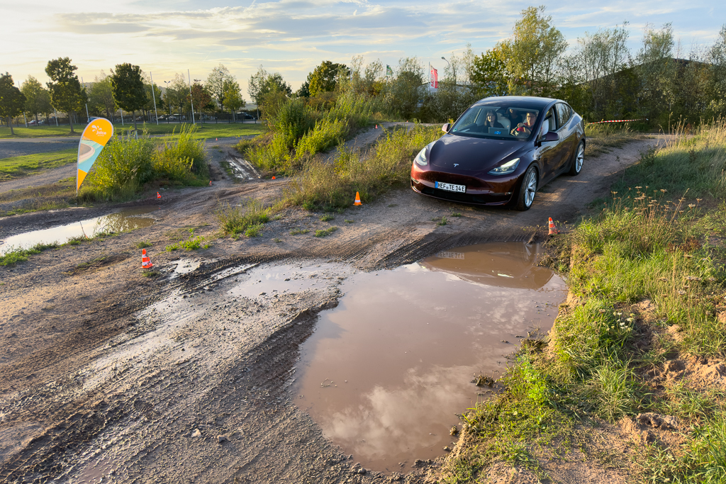 A Tesla Model 3 navigating a muddy terrain with puddles, surrounded by greenery and flags in the background.