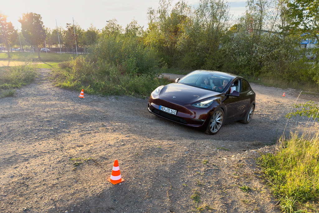A purple Tesla Model Y navigating through a rough, gravel path marked with traffic cones, surrounded by greenery and trees, during sunset.