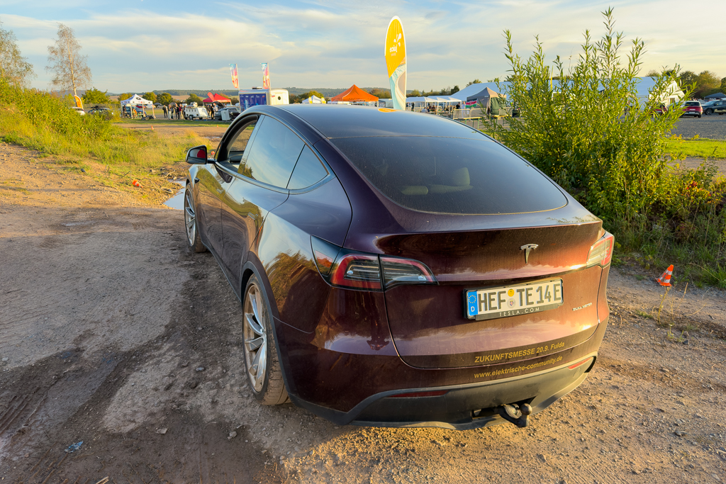A maroon Tesla Model 3 parked on a dirt road with green grass and trees in the background, featuring tents and vehicles in the distance under a partly cloudy sky.