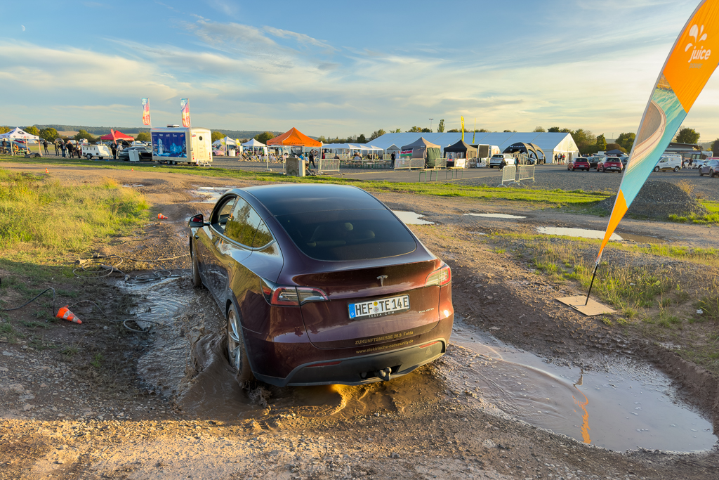 A maroon Tesla vehicle navigating through a muddy area at an outdoor event, with festival tents and people in the background.