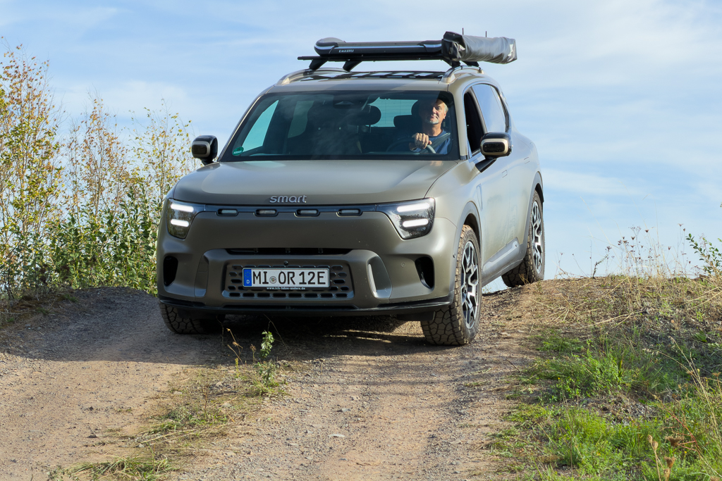 A smart electric SUV navigating a gravel path, with a driver visible inside. The vehicle features a rooftop tent and alloy wheels, surrounded by greenery and a blue sky.