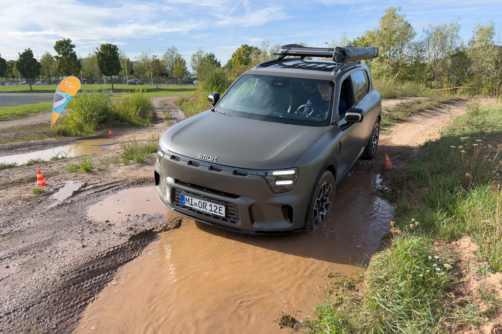 A matte grey smart vehicle navigating through a muddy puddle on an off-road track, with a driver visible inside. In the background, there are trees and a colorful banner.