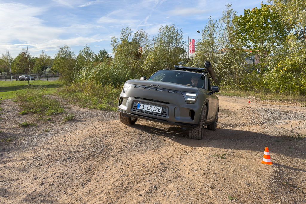 A rugged vehicle navigating a dirt path surrounded by greenery, with traffic cones marking an obstacle course in the background.