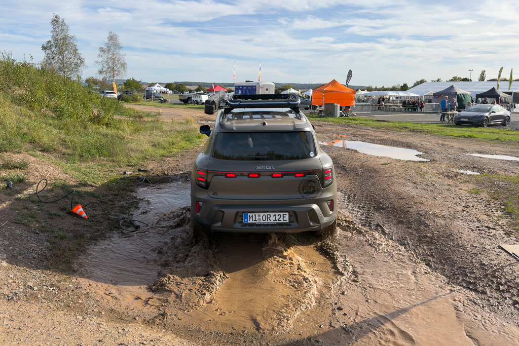 A vehicle driving through a muddy puddle in an outdoor event area, surrounded by tents and grassy terrain under a partly cloudy sky.