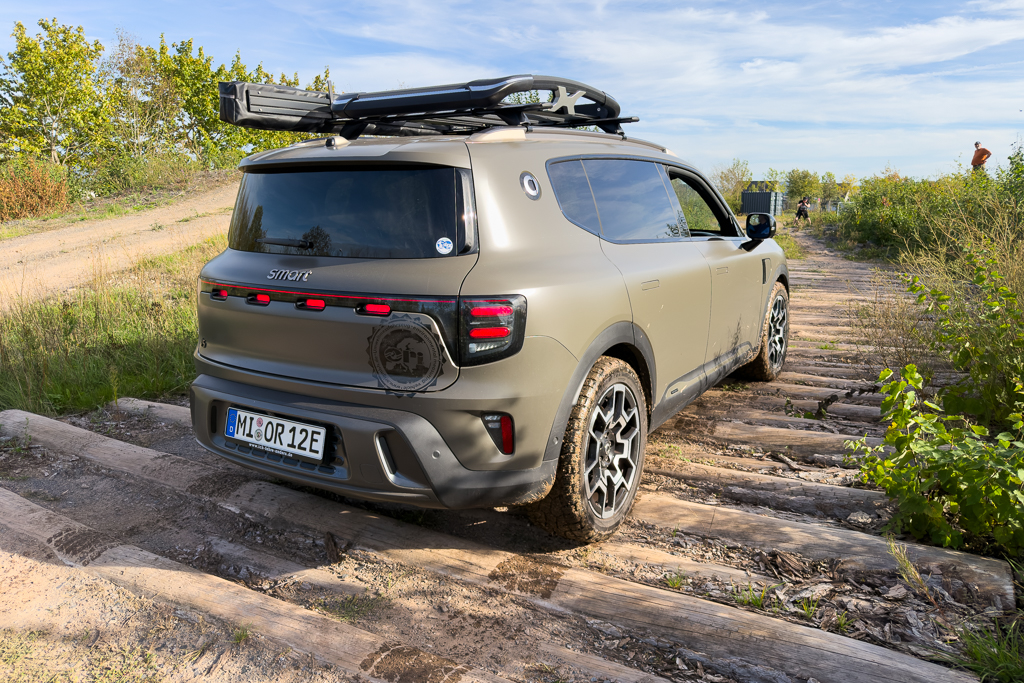 A smart SUV parked on a rugged dirt path surrounded by greenery, featuring a roof rack and off-road tires.