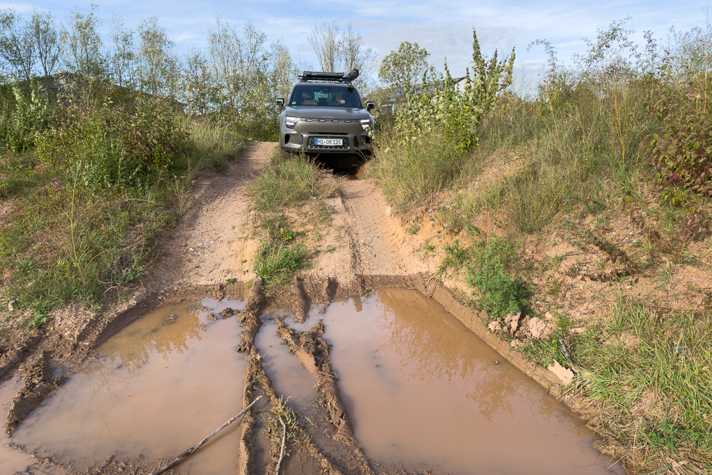 An off-road vehicle positioned on a rugged dirt path with muddy patches and surrounding vegetation under a clear sky.