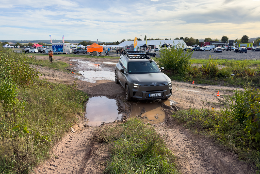 A small SUV navigating through a muddy trail with puddles, surrounded by greenery and tents in the background.