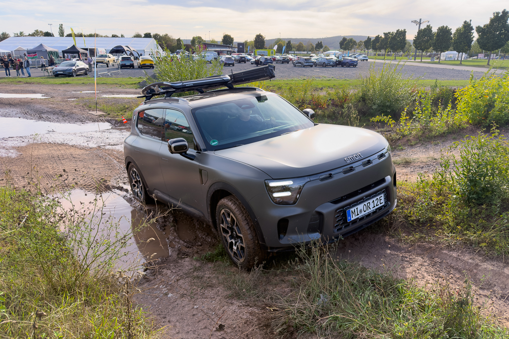 A gray compact car with a roof rack navigating through muddy terrain, surrounded by green grass and a cloudy sky in the background.
