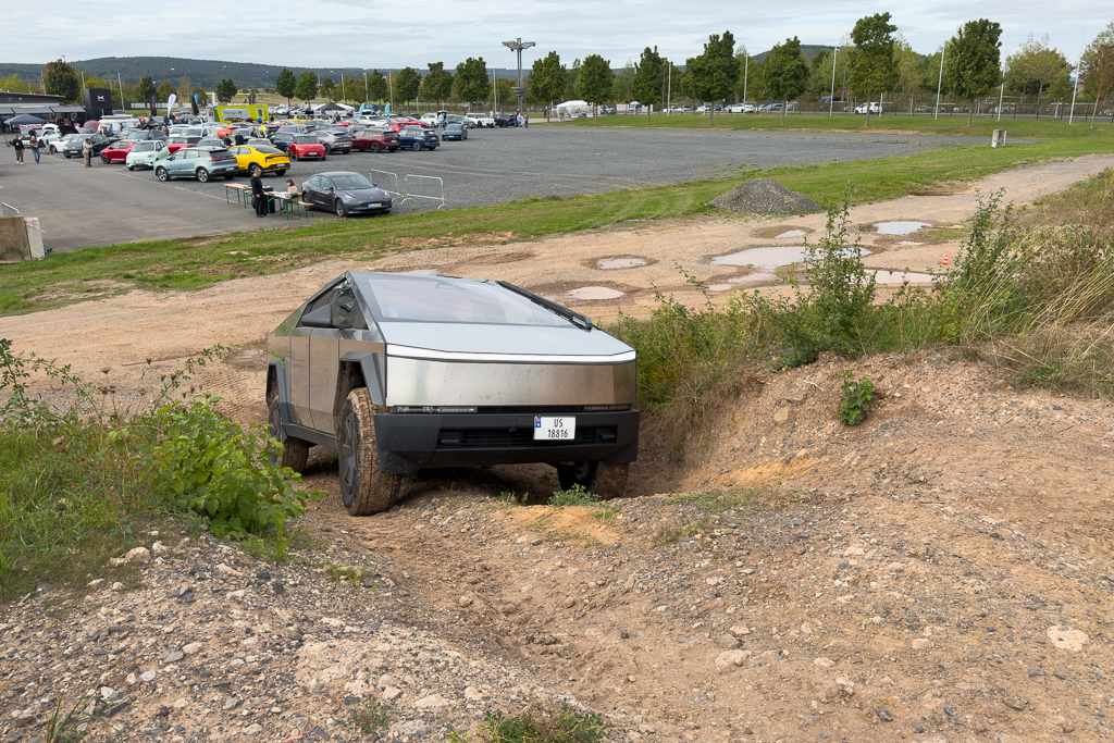 A Tesla Cybertruck navigating a rugged dirt path on an outdoor test track with other parked vehicles in the background.