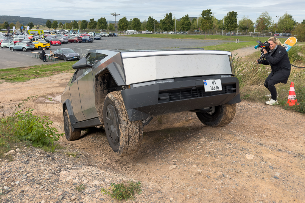 A futuristic vehicle, possibly a concept truck, navigating an uneven, muddy terrain while a woman films it with a camera. In the background, other parked cars and trees are visible.