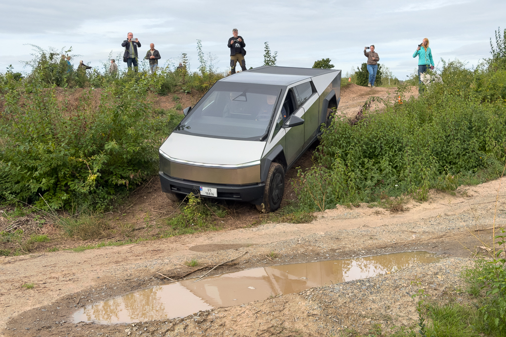 A futuristic electric vehicle parked on uneven terrain surrounded by tall grass and bushes, with several people in the background observing and taking photos.