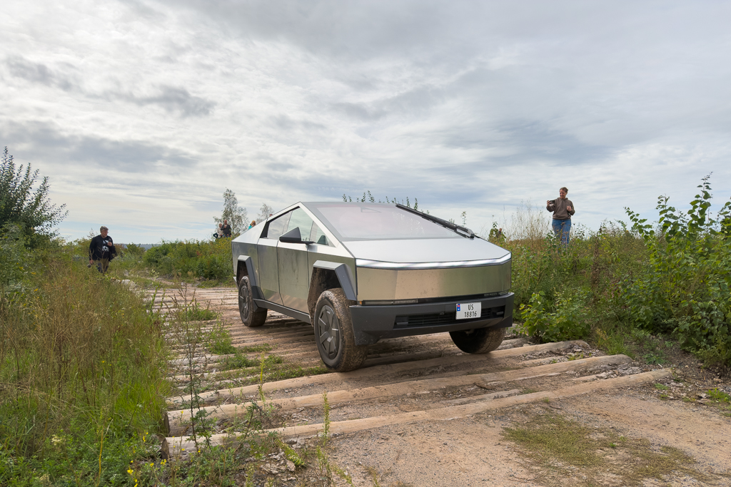 A futuristic electric vehicle with a distinctive angular design, driving over wooden planks on a rugged terrain, with two people in the background observing.