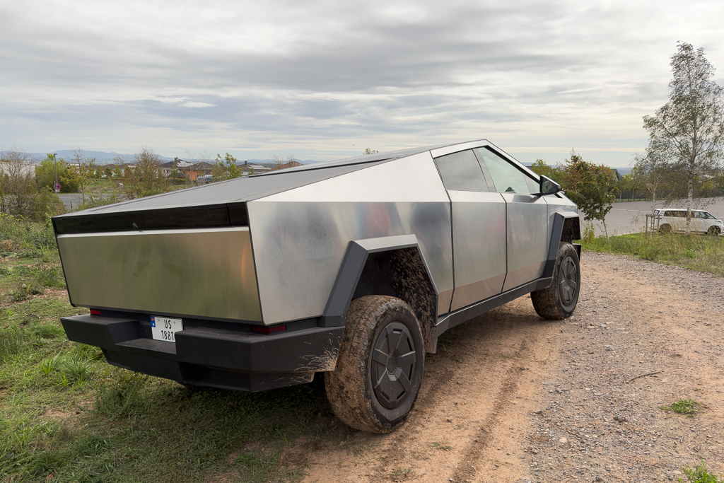 A futuristic, angular electric vehicle with a metallic exterior, positioned on a grassy dirt path with cloudy skies in the background.