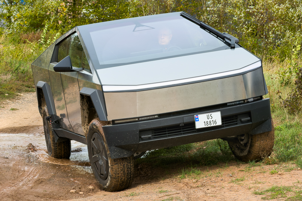 A futuristic, metallic electric truck is driving on a dirt path surrounded by greenery, showcasing its rugged design and off-road capability.