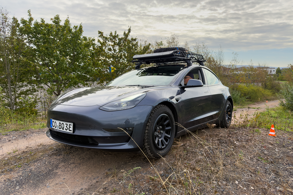A gray electric car parked on a gravel path, equipped with off-road tires and a roof rack, surrounded by greenery and trees.