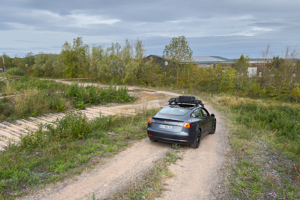 A gray car with a roof rack driving on a gravel path surrounded by greenery and trees under a cloudy sky.