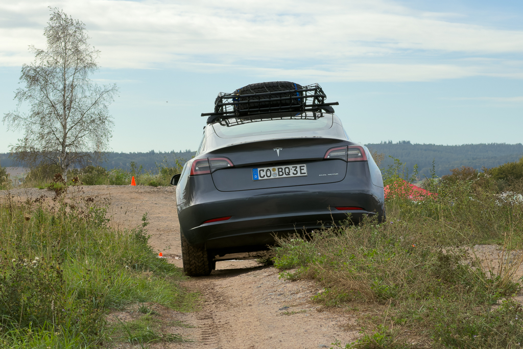 A Tesla car parked on a gravel path surrounded by tall grass and trees, with a roof rack on top. Scenic landscape in the background.