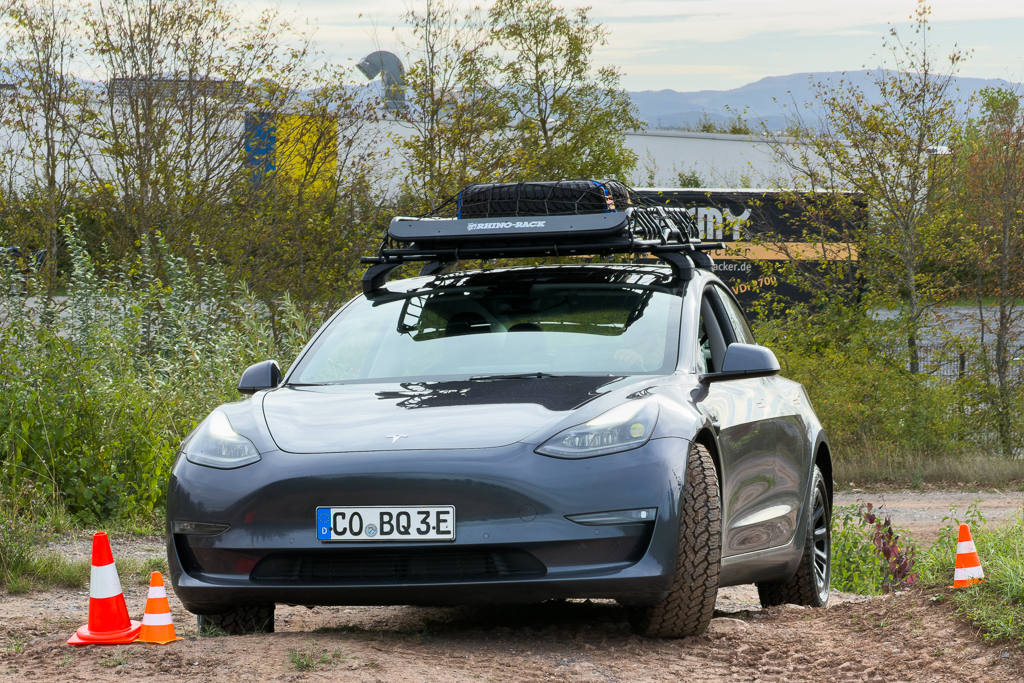 A gray electric SUV parked on a gravel road with cones nearby, featuring a roof rack.