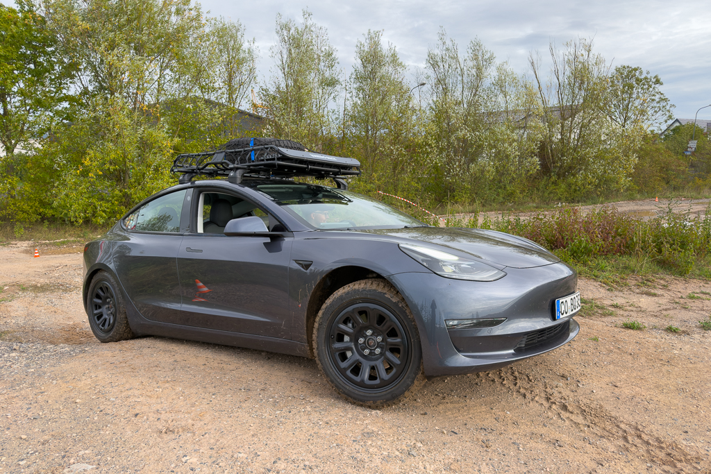 A gray Tesla Model 3 parked on a dirt road, featuring a roof rack and surrounded by greenery.