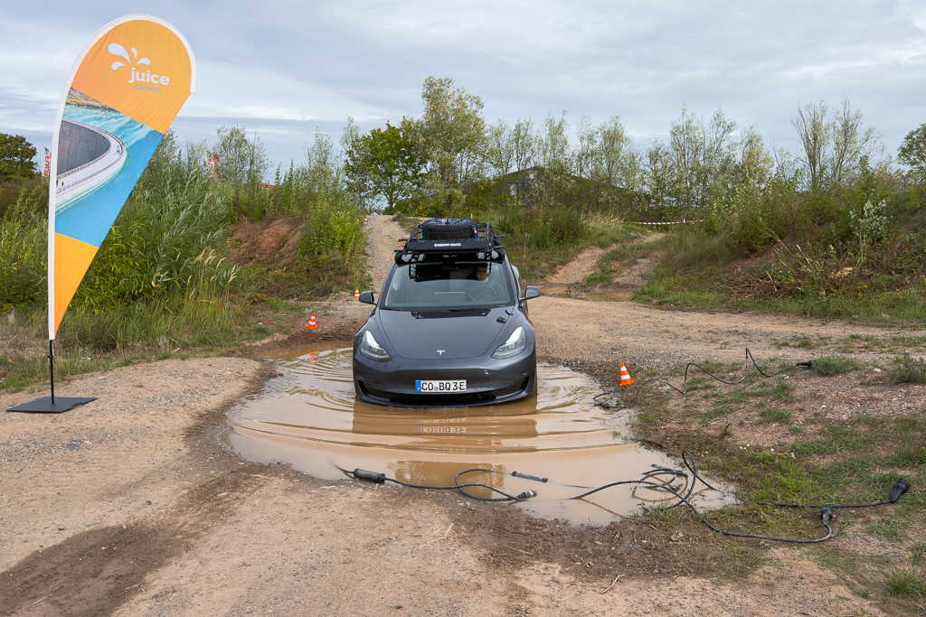 A Tesla electric vehicle partially submerged in a puddle on a dirt path, with a power supply cable visible nearby. A Juice Power flag is positioned on the left, indicating charging availability.