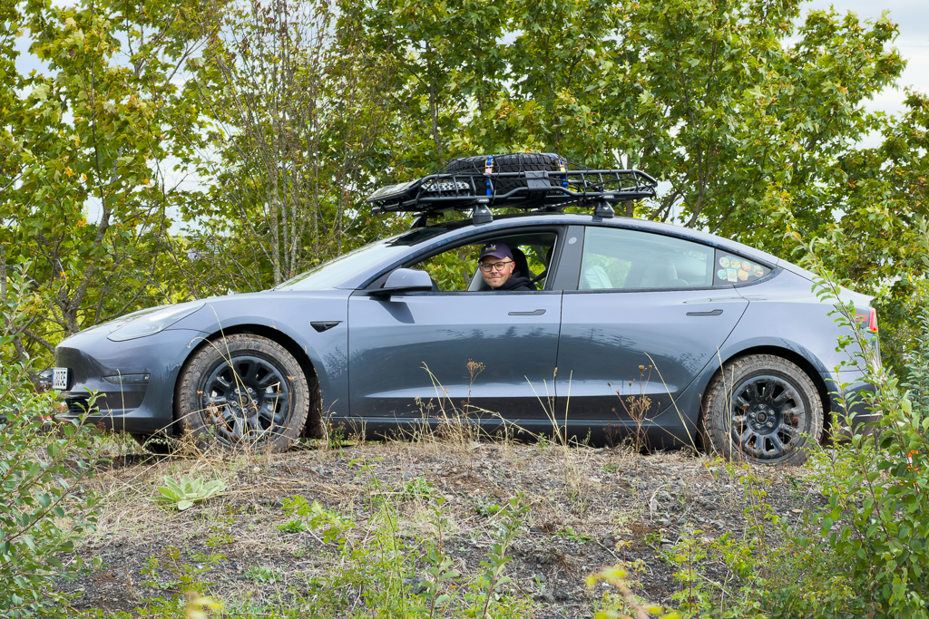 A person inside a gray electric car parked on a gravel hill, surrounded by greenery and trees, with a roof rack equipped for outdoor activities.