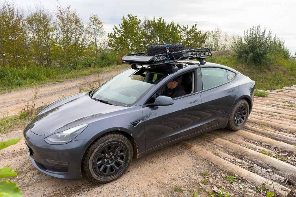 A gray Tesla Model 3 parked off-road with a rooftop cargo carrier, surrounded by greenery and dirt paths.