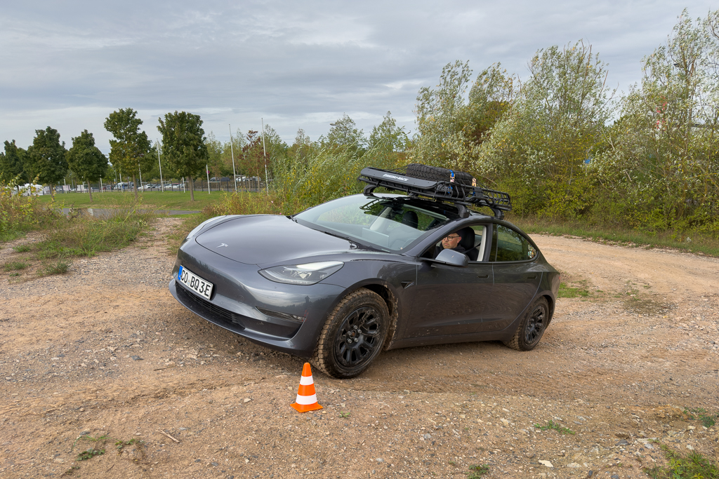 A Tesla Model 3 parked on a dirt path, with a person inside, showcasing an off-road setup with a roof rack and spare tire.