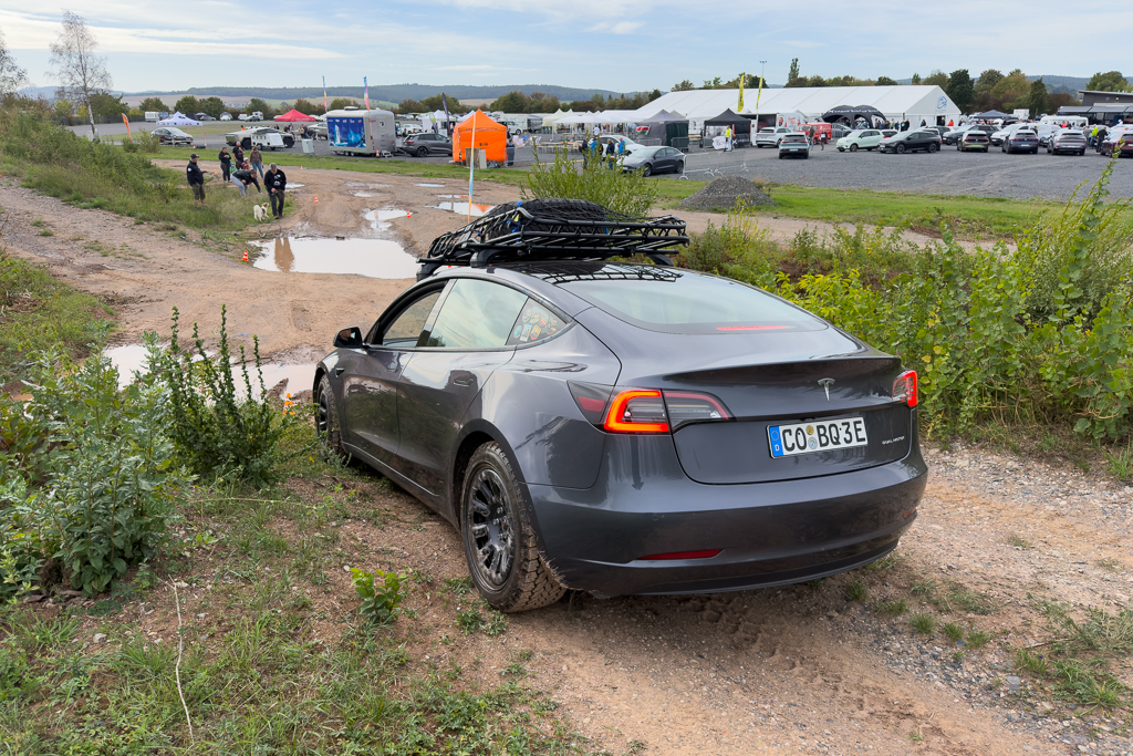 A gray Tesla car with a roof rack parked near a muddy off-road path, surrounded by green vegetation and people in the background at an event.