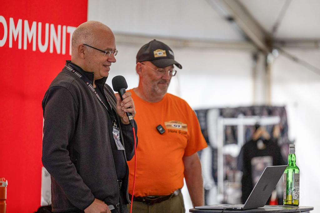 Two men speaking at a presentation, one holding a microphone and the other standing beside a laptop on a table, with a red backdrop featuring the word 'COMMUNITY'.