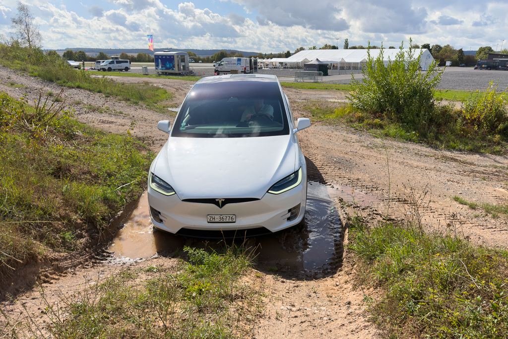 A white Tesla Model X navigating through a muddy area with puddles, surrounded by grass and dirt, with a distant view of parked vehicles and tents in the background.