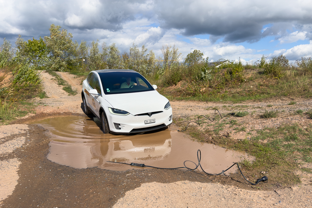 A white electric vehicle driving through a puddle on a gravel road, with charging cables visible on the ground, surrounded by greenery and cloudy skies.